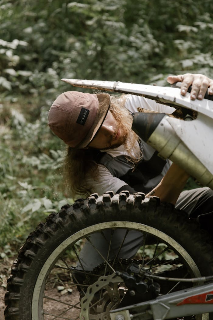 A man focuses intently as he repairs a motorcycle in a lush outdoor setting.