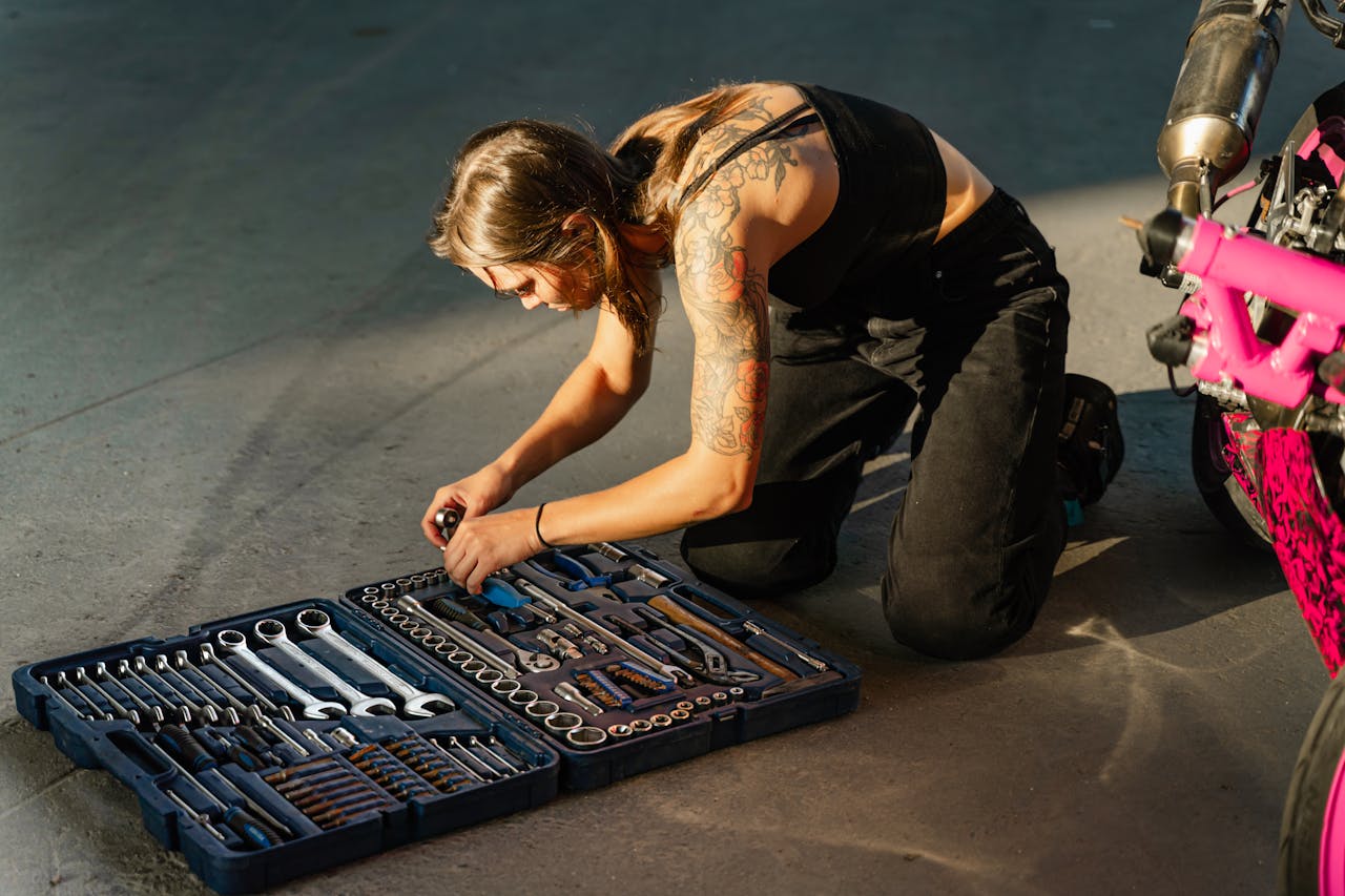 Young tattooed woman in black tank top organizing tools from a toolbox beside a motorcycle.